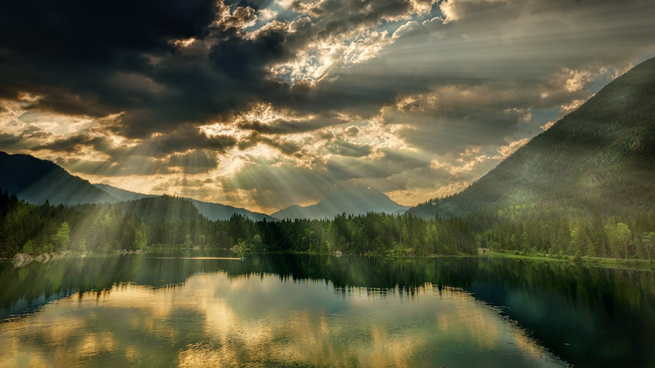 A stunning view of a tranquil lake surrounded by mountains with sunrays piercing through clouds.